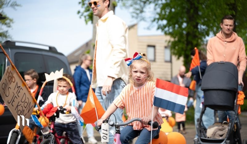 Child wearing orange striped shirt rides a small bicycle in a parade, holding a Dutch flag with orange decorations nearby and onlookers in the background.
