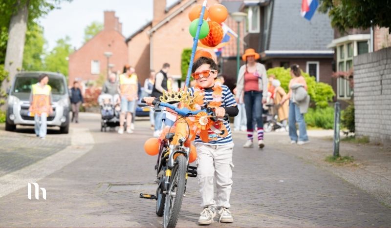 Child wearing orange sunglasses rides a small bike decorated with balloons down a residential street parade, with spectators in the background.