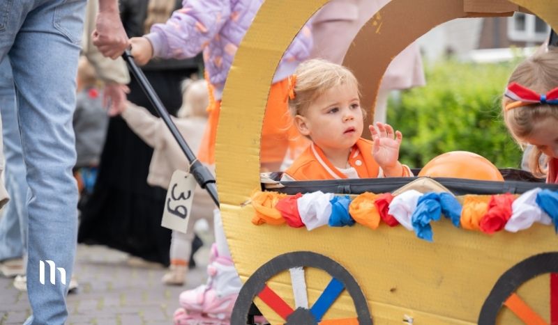 Young child in orange, sitting in a colorful homemade parade cart with garland, waving hand.