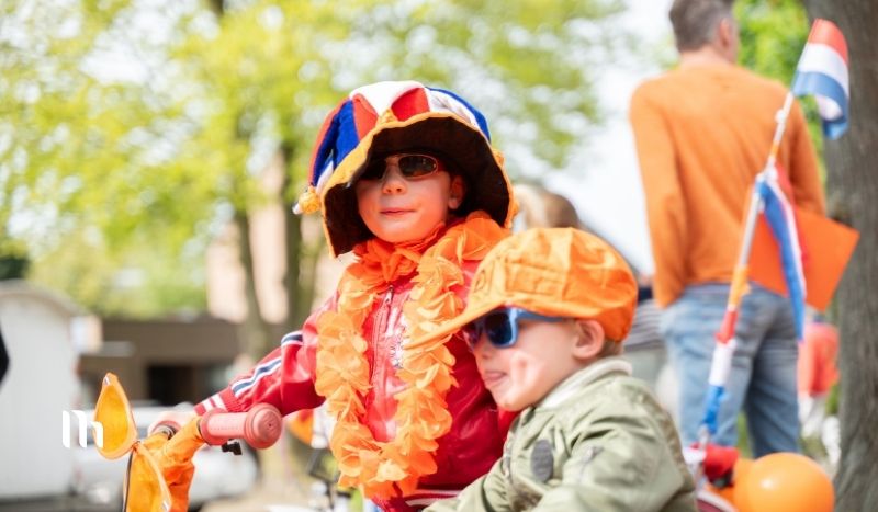 Two children in bright costumes riding bicycles at a parade, orange boas and hats, Dutch flag visible in the background; sunny day.