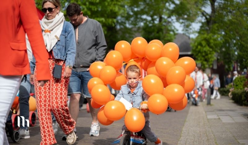 Young child on a small bike rides through a street parade, surrounded by a bright ring of orange balloons.