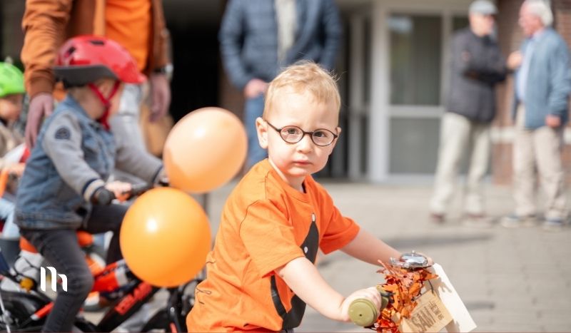 A young child wearing an orange shirt and glasses rides a scooter at an outdoor event, with orange balloons and other kids in the background.