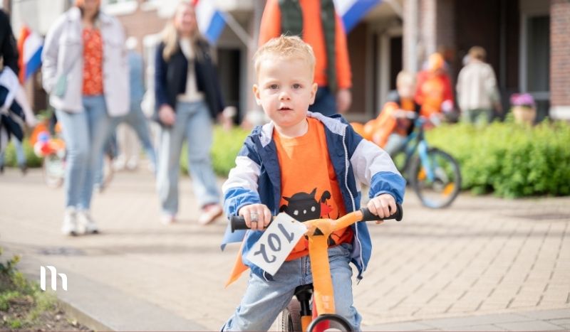 Child riding a small bike in a street parade, wearing an orange shirt and blue jacket, with race number 701 attached to the handlebars.
