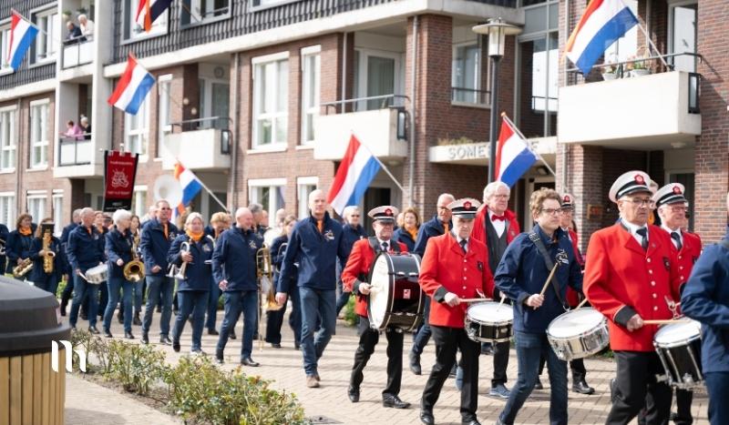 Street parade with a marching band performing along a brick apartment row; front-line drummers wear red jackets and carry drums, with Dutch flags overhead.