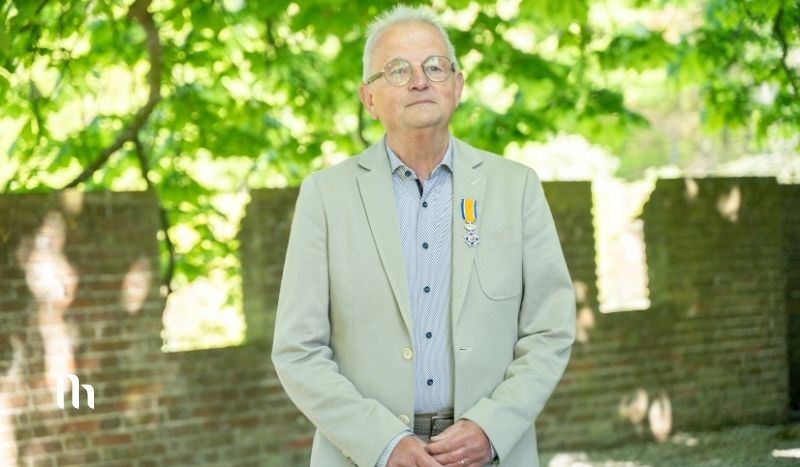 Portrait of a senior man in a light gray blazer with a medal on his chest, standing outdoors with a brick wall and green trees behind him.