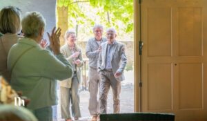 Group of elderly people smiling and clapping as they enter through a sunlit doorway into a room inside, welcoming atmosphere.