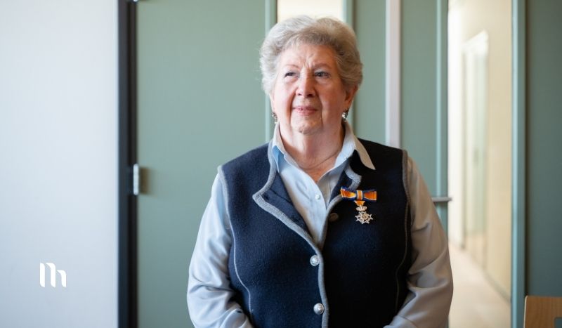 Older woman with short gray hair wearing a navy vest over a light blue shirt, medals on her chest, standing in a hallway.