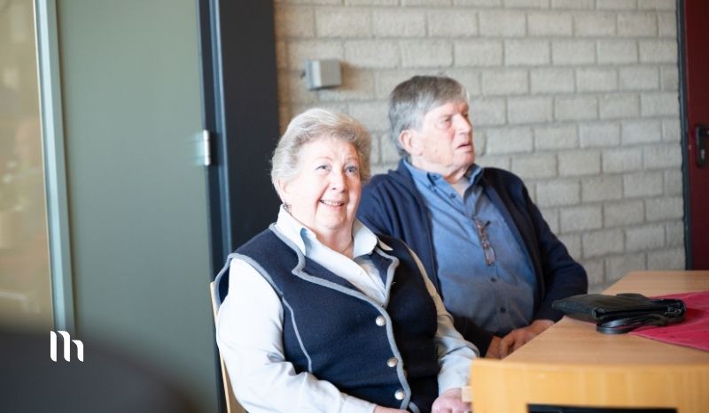 Two older adults sit at a table in a brick-walled room; the woman in a navy vest smiles at the camera, the man looks to the side.