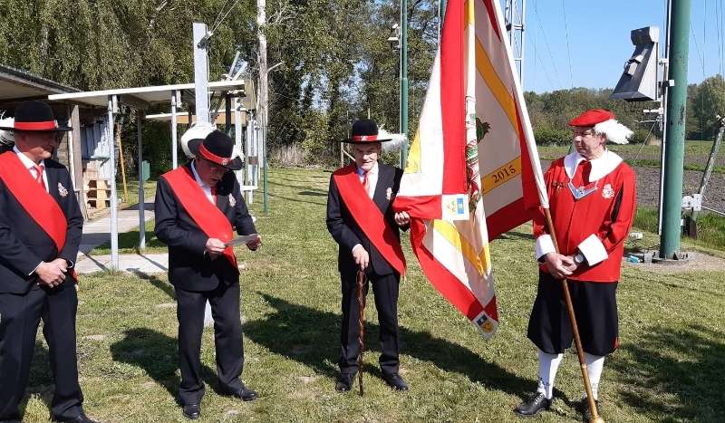 Four men in formal uniforms with red sashes standing outdoors, one holding a ceremonial flag.