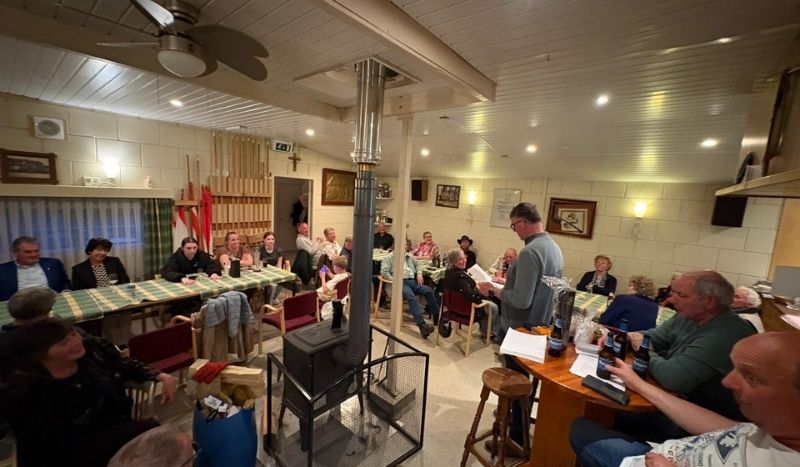 Community gathering around tables in a cozy room with a central wood stove; a man stands addressing the group.