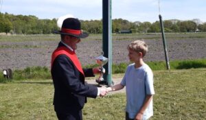 Older man in a suit and hat hands a trophy to a smiling boy while shaking hands outdoors on a grassy field.