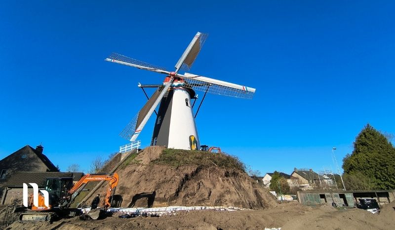 Windmill under construction atop a dirt hill, with a backhoe digging at the site and a clear blue sky behind.