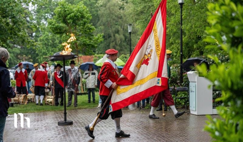 Group of people in historical costumes marching with a large red, white, and gold flag in a park ceremony; a flaming brazier and onlookers with umbrellas nearby.