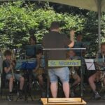 Youth concert band under a canopy, conductor guiding the players while children read sheet music, outdoors in a park; banner reads 'St. Nicasius'.