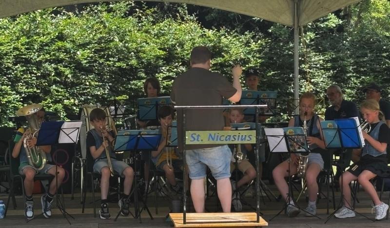 Youth concert band under a canopy, conductor guiding the players while children read sheet music, outdoors in a park; banner reads 'St. Nicasius'.