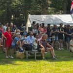 Large crowd of people standing and sitting on benches in a park, watching an event near a white tent with flags.