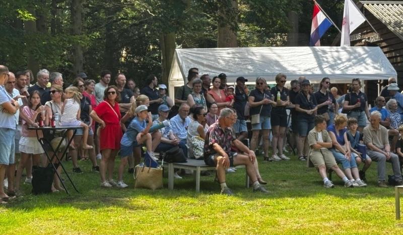 Large crowd of people standing and sitting on benches in a park, watching an event near a white tent with flags.