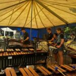 Group of young percussionists performing under a large yellow tent, playing marimba, xylophones, and drums outdoors.