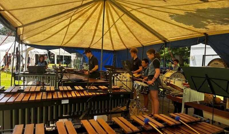 Group of young percussionists performing under a large yellow tent, playing marimba, xylophones, and drums outdoors.
