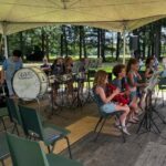 Youth band under a beige canopy performing in a park, with children playing brass instruments and drums and sheet music stands; empty green chairs surround the wooden stage.