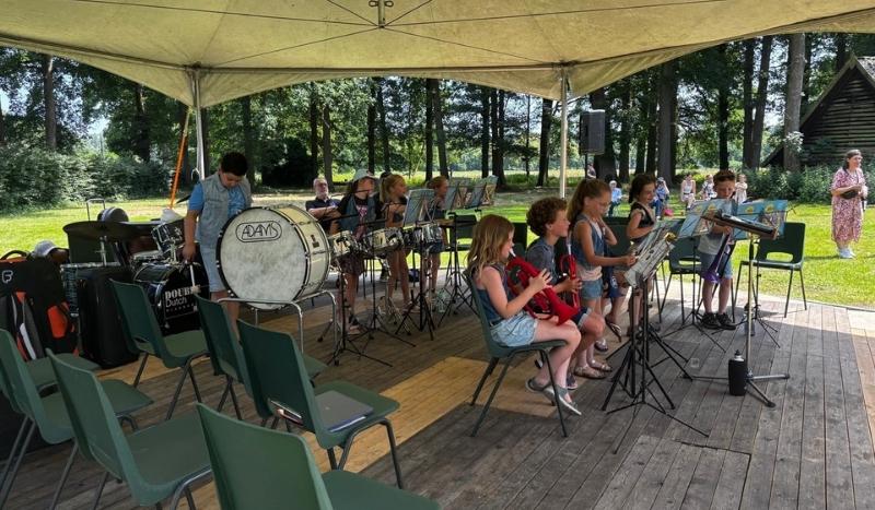 Youth band under a beige canopy performing in a park, with children playing brass instruments and drums and sheet music stands; empty green chairs surround the wooden stage.