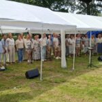 Outdoor choir performs under a white canopy; a conductor faces the group from the right, with a colorful banner on the left.