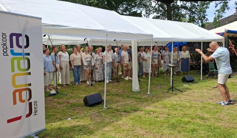 Outdoor choir performs under a white canopy; a conductor faces the group from the right, with a colorful banner on the left.