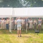 Outdoor choir under white tents with a conductor facing the singers; a group of casually dressed people singing outdoors on a grassy lawn.