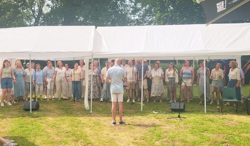 Outdoor choir under white tents with a conductor facing the singers; a group of casually dressed people singing outdoors on a grassy lawn.