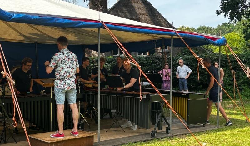 A group of musicians playing steel drums under a blue-and-white canopy in an outdoor venue, with a conductor leading from a wooden platform.