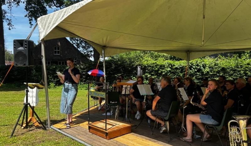 Woman singing into a microphone on an outdoor stage under a beige canopy, with a seated band and music stands behind her.