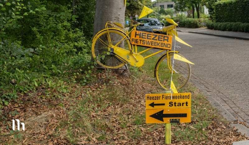 A bright yellow bicycle mounted against a tree with signs for 'Heezer Fiets Weekend' and a separate yellow 'Start' sign with left-pointing arrows on a street corner.