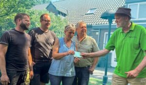 A man in a green polo hands a check to a woman while four others smile and watch in a sunny garden.