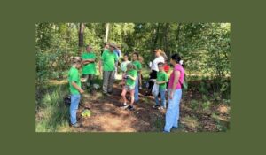 Group of children and adults in a forest clearing, some wearing matching green shirts, gathered for an outdoor activity.
