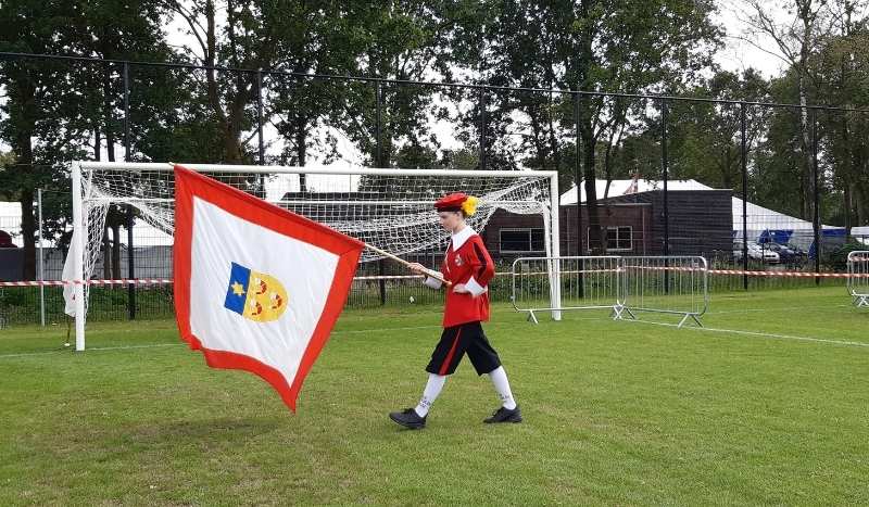 Flag bearer: a child in a red outfit marching on a grassy field, carrying a large white flag with a red border and a blue shield emblem, near a soccer goal during a parade or event.