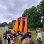 Children gather on a grassy field around a large orange and purple inflatable slide while adults supervise.