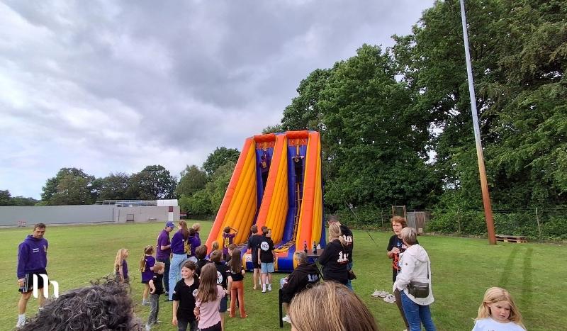 Children gather on a grassy field around a large orange and purple inflatable slide while adults supervise.