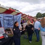 Outdoor community fair with red-and-white striped tents, banners, and people chatting at information booths on a grassy field.