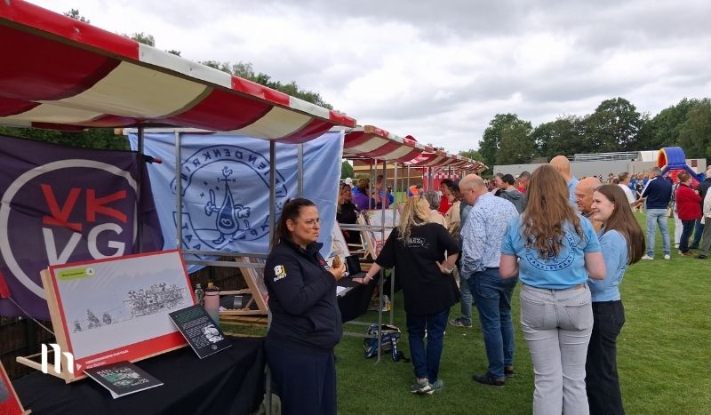 Outdoor community fair with red-and-white striped tents, banners, and people chatting at information booths on a grassy field.