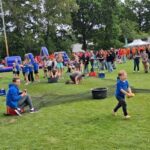 Children in blue shirts play games on a grassy field at an outdoor festival, with inflatables and a crowd in the background.