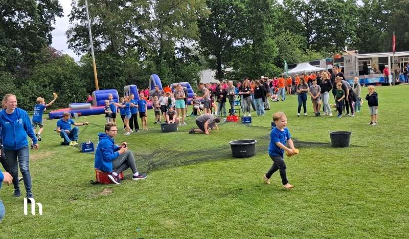 Children in blue shirts play games on a grassy field at an outdoor festival, with inflatables and a crowd in the background.