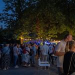 A crowd of people mingling at an outdoor evening event under string lights, with trees and a stage in the background.