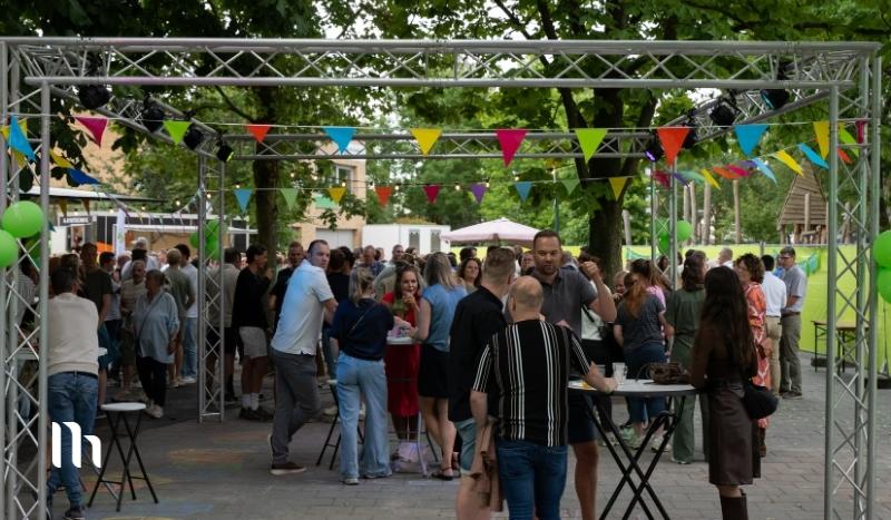 Outdoor community event under a metal truss with colorful pennant bunting; crowd mingling around high tables in a park.