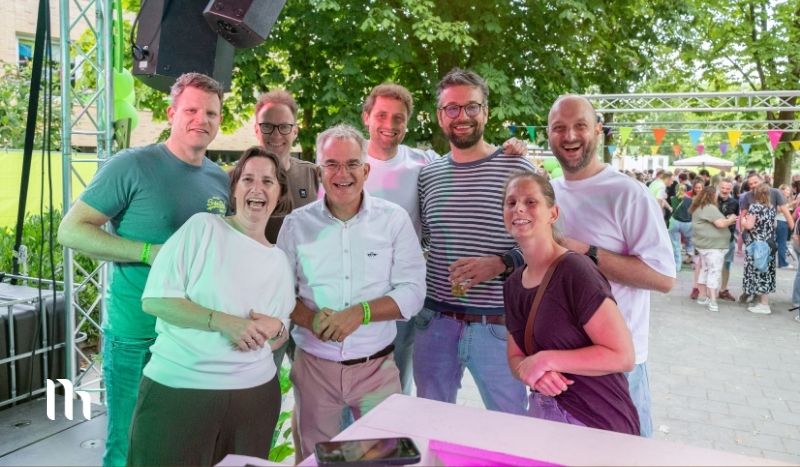 Eight adults smile for a group photo at an outdoor festival stage with colorful bunting and a crowd behind them.