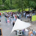 Outdoor community festival scene with a crowd gathered around tents and stalls, surrounded by bright lime-green fencing and trees.