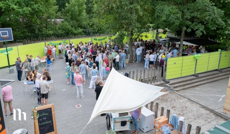 Outdoor community festival scene with a crowd gathered around tents and stalls, surrounded by bright lime-green fencing and trees.