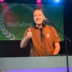 Smiling man in a brown shirt stands at a stage podium with headphones, giving a thumbs up under colorful stage lights, a green banner with a laurel wreath behind him.