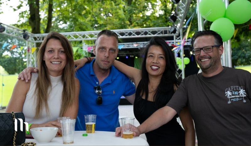 Four friends posing together at an outdoor event, standing behind a white high-top table with drinks, smiling with arms around each other; stage and green balloons visible in a park-like setting.