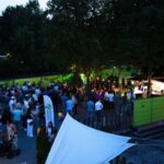Evening outdoor festival with a crowd gathering around food stalls under trees and green barriers nearby.