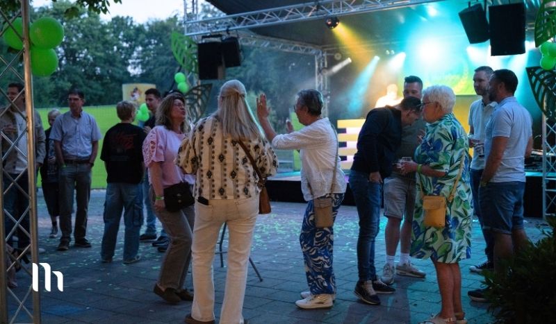Group of people chatting near an outdoor stage with green balloons and blue/green lighting at dusk in a park setting